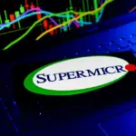 Smartphone showing the Supermicro logo on a laptop keyboard in front of a stock price candlestick chart