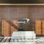 Reception desk in front of a wood-panelled wall with the Capital One logo in a bright office lobby, with people walking through
