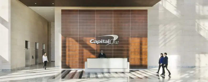 Reception desk in front of a wood-panelled wall with the Capital One logo in a bright office lobby, with people walking through