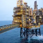 Offshore gas production platform with illuminated yellow topsides standing in open sea at dusk