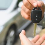 A hand holding out a car key fob above an open palm in front of a blurred car