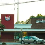 Wendy's restaurant exterior with the red logo tower and white Wendy's sign on a brick wall, with a car parked in front