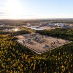 Aerial view of an oil sands operation, with a cleared industrial well pad and a distant processing plant surrounded by autumn boreal forest