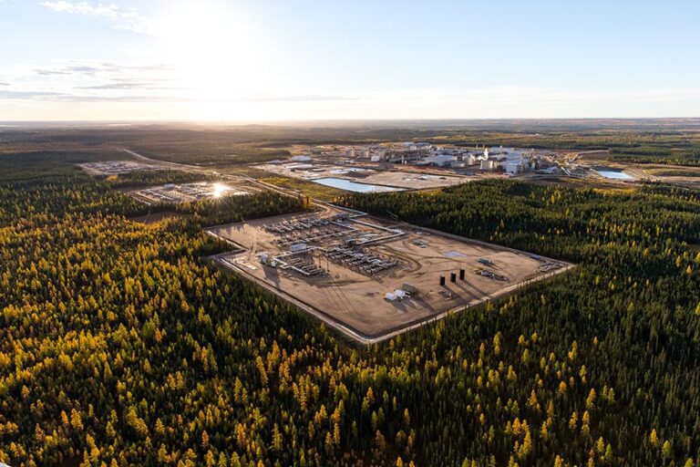 Aerial view of an oil sands operation, with a cleared industrial well pad and a distant processing plant surrounded by autumn boreal forest