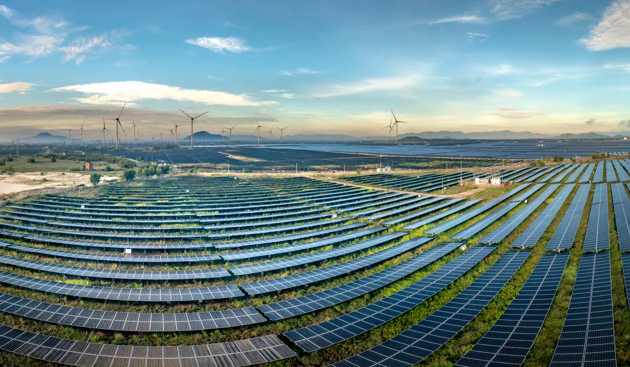 Aerial view of a large solar farm with rows of panels in the foreground and wind turbines along the horizon under a blue sky