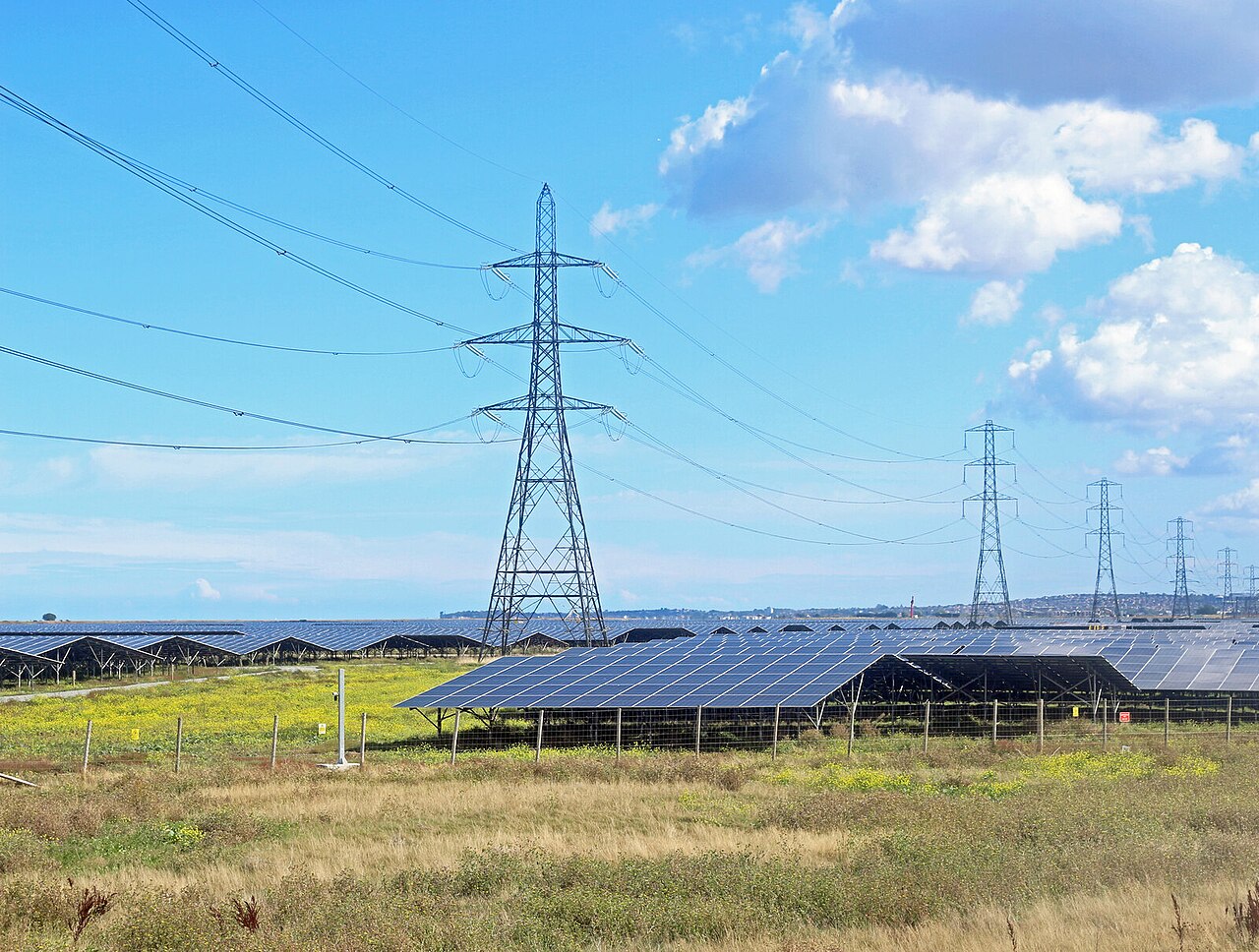 Rows of solar panels on open land with tall steel transmission pylons and power lines under a blue sky